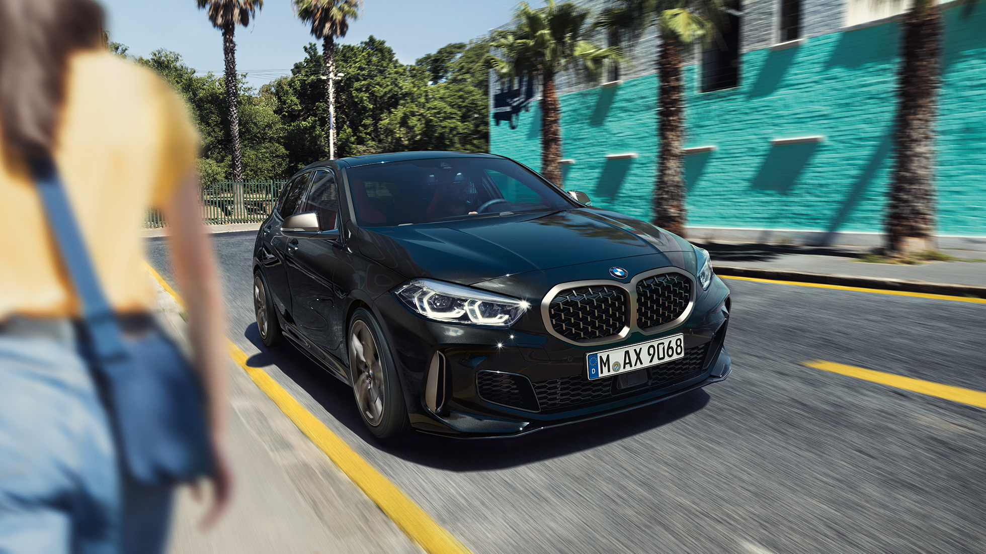 BMW M135i xDrive, three-quarter front view. BMW M135i xDrive in Black Sapphire metallic, three-quarter front view driving, with woman in foreground.