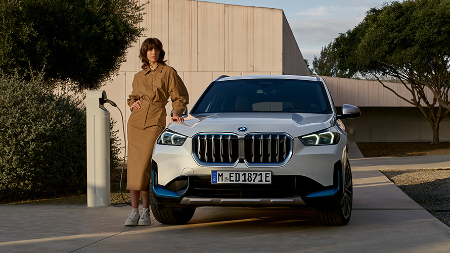Woman in brown dress leaning against a BMW iX1 U11 2022 front view next to a charging station