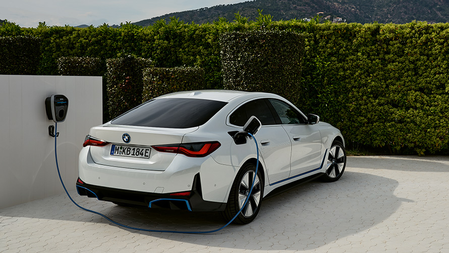 Charging at the BMW Wallbox Gen 3 BMW i4 G26 BEV Three-quarter rear view  BMW i4 G26 BEV three-quarter rear view charging connected to BMW Wallbox Gen 3 in front of hedge
