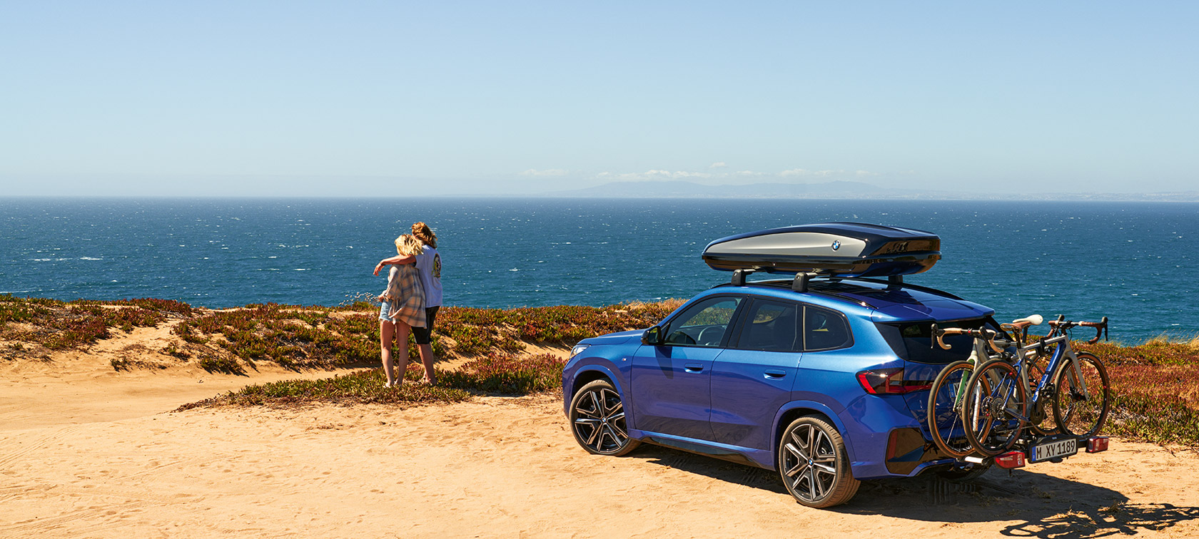 Original BMW Accessories for Spring and Summer BMW X1 U11 Original BMW Accessories for Spring and Summer BMW X1 U11 standing on sand in front of a coast with 2 people